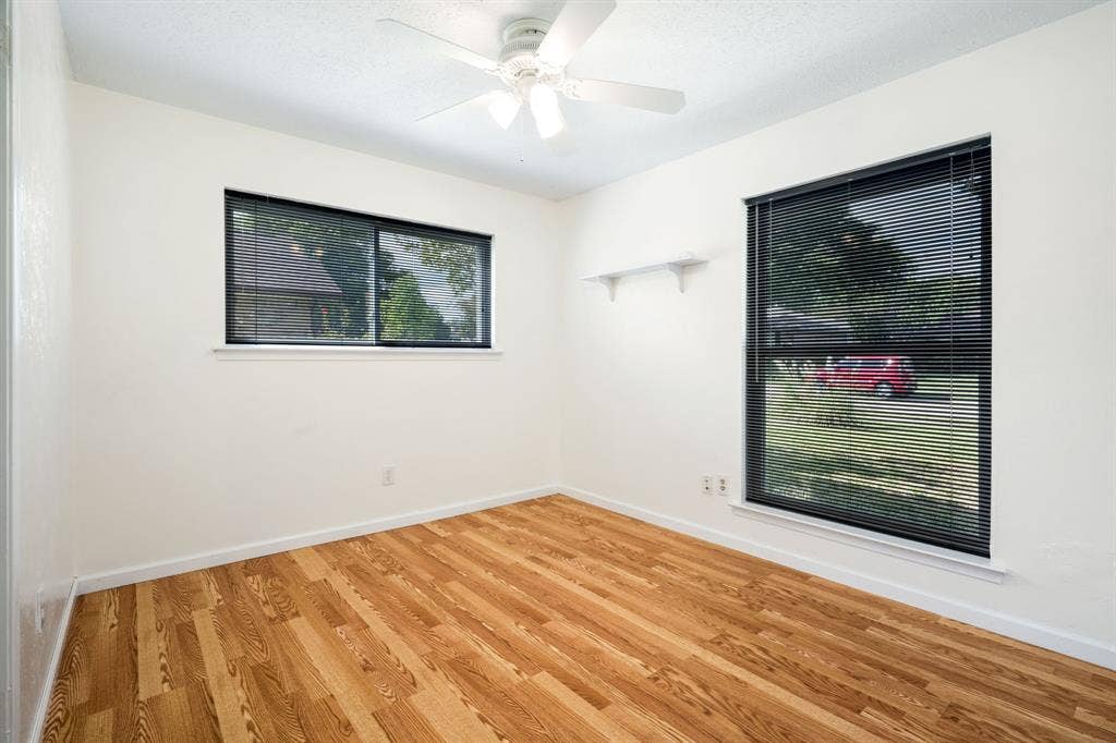 Room featuring wood-finish flooring, a white ceiling fan with integrated lighting, dual windows with horizontal blinds, and a built-in wall-mounted shelf