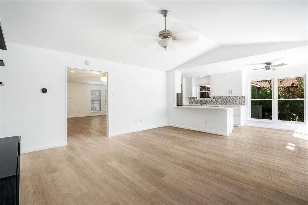 Open-concept floor plan featuring vaulted ceilings, wood-finish flooring, a kitchen with white cabinetry, a stone-tile backsplash, and a breakfast bar