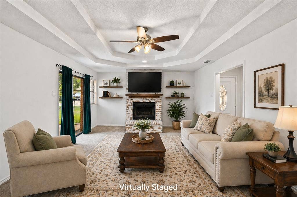 Living area featuring a white-painted brick fireplace with a wood mantel, tray ceiling, ceiling fan, and sliding glass door