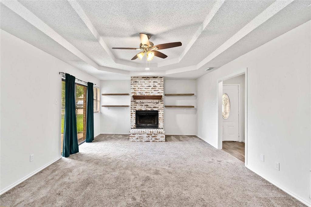 Living area featuring a white brick fireplace with wood mantel and open shelving, gray carpet, a tray ceiling with a ceiling fan, and a sliding glass door