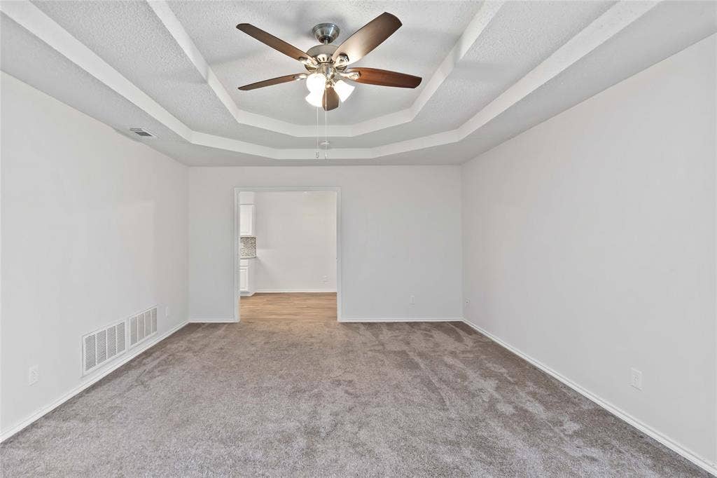 Recessed tray ceiling with a ceiling fan, plush gray carpeting, and white baseboards