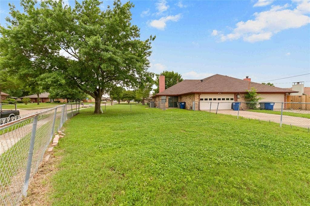 Expansive lawn area featuring a mature tree, chain-link fencing, and a paved driveway offering access to the attached two-car garage