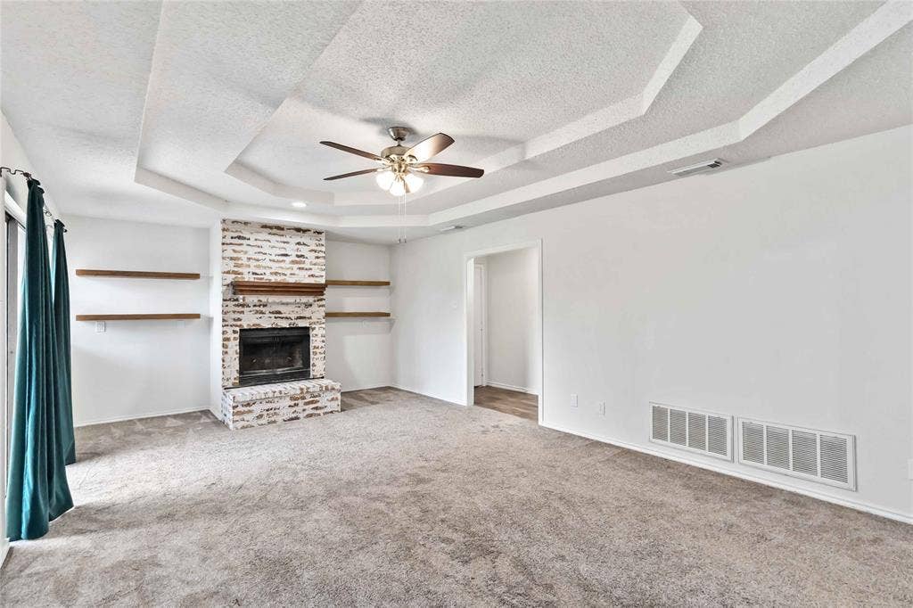 Spacious living area featuring a tray ceiling, recessed fireplace with a brick surround, and built-in floating shelves