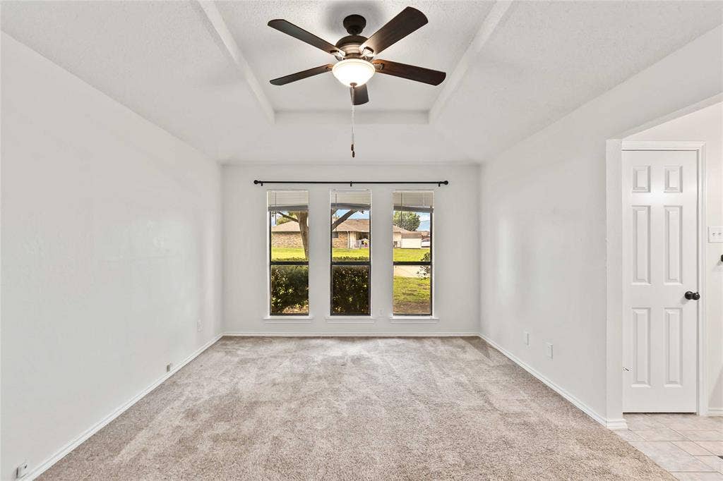 Bright room featuring a tray ceiling, neutral-toned carpeting, a ceiling fan with integrated lighting, and three tall windows