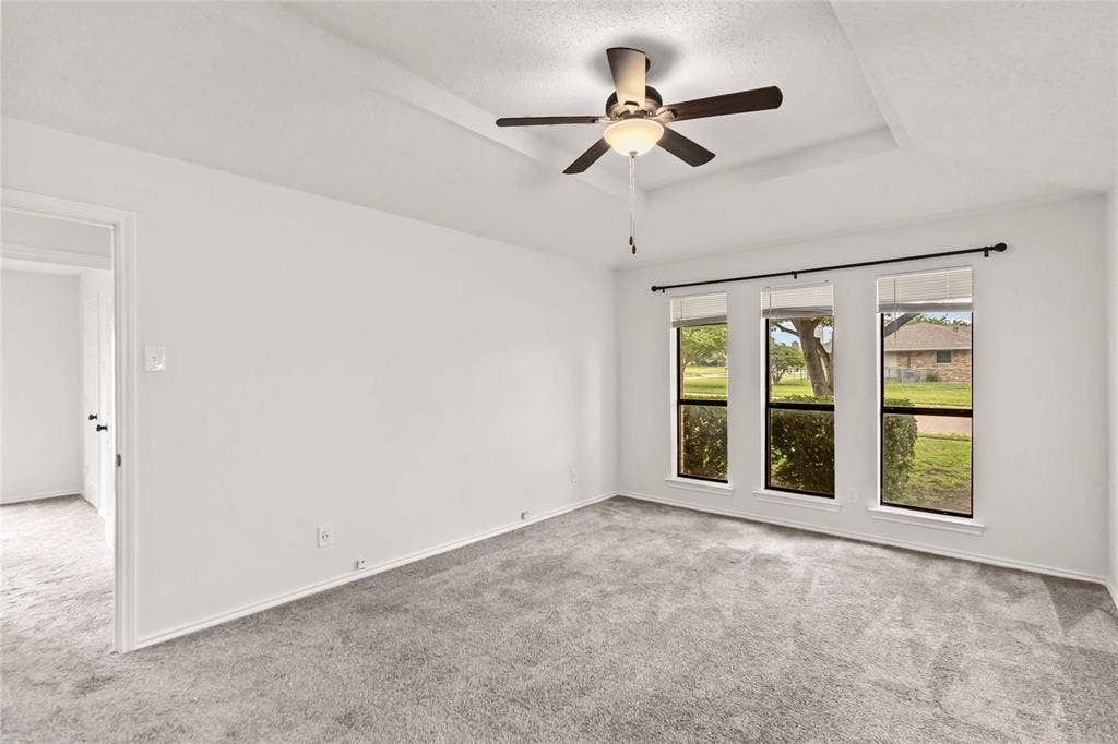 Room featuring light gray carpet, three windows with dark frames, a tray ceiling, a ceiling fan with light, and white walls