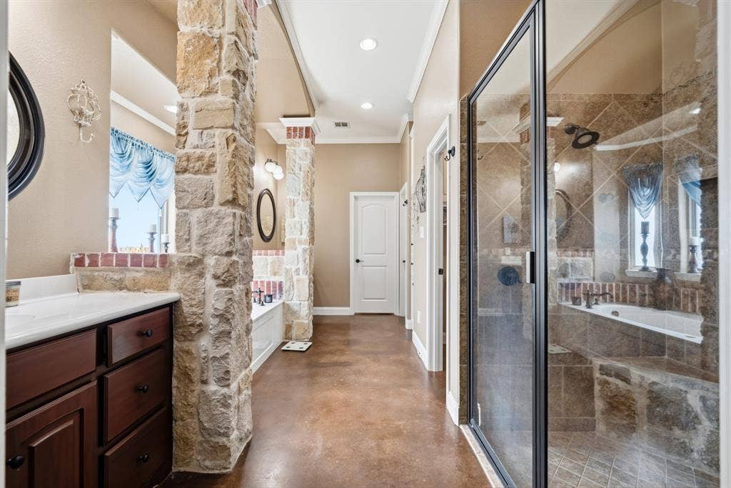 1021 Cascade Trail, McGregor, TX 76657 Walk-in shower with glass enclosure, soaking tub with tile surround, stone-clad architectural columns, dark wood-finish vanity with white countertop, and concrete-finish flooring