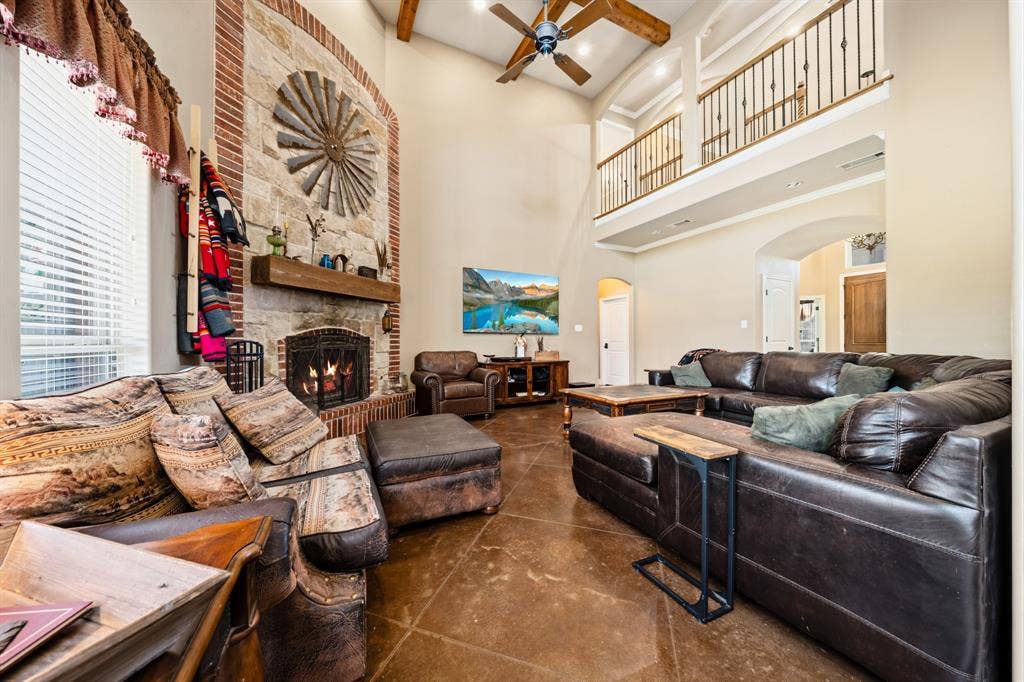 1021 Cascade Trail, McGregor, TX 76657 Vaulted ceiling with exposed wood beams, stone fireplace with a wood mantle, upper-level balcony with metal spindle railing, and stained concrete flooring
