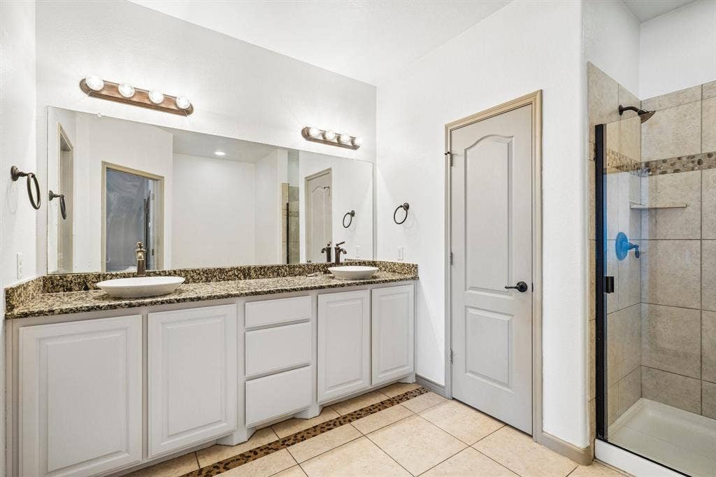 7937 Parkwood Boulevard, Plano, TX 75024 Dual vessel sinks, granite-finish countertop, white cabinetry with bronze hardware, tiled shower with glass enclosure, and neutral tone tile flooring
