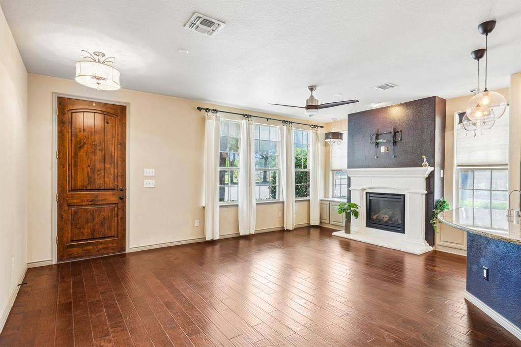 7937 Parkwood Boulevard, Plano, TX 75024 Rich wood-finish flooring, a prominent fireplace with a white mantel, multiple windows with white trim, a decorative entry door, and a built-in ceiling fan with lighting