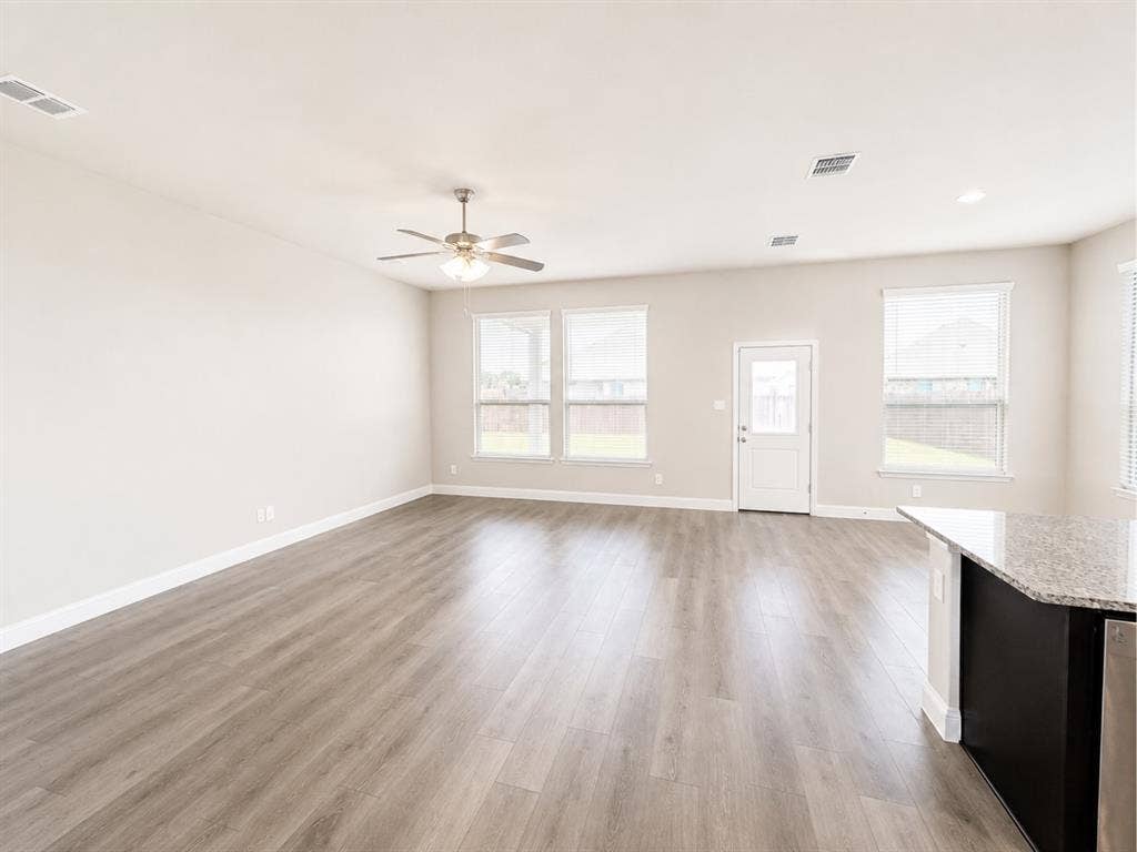 6002 14th Street, Midlothian, TX 76065 Spacious living area featuring light wood-finish flooring, a ceiling fan with integrated lighting, multiple windows with blinds, a white door providing exterior access, and a kitchen island with a granite countertop