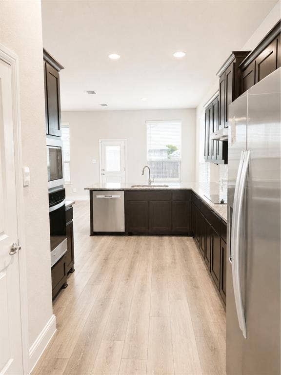 6002 14th Street, Midlothian, TX 76065 Modern galley kitchen featuring dark cabinetry with integrated hardware, stainless steel appliances including a side-by-side refrigerator and built-in microwave, a contrasting dark island with undermount sink and prep space, recessed ceiling lighting, and light wood-finish flooring throughout