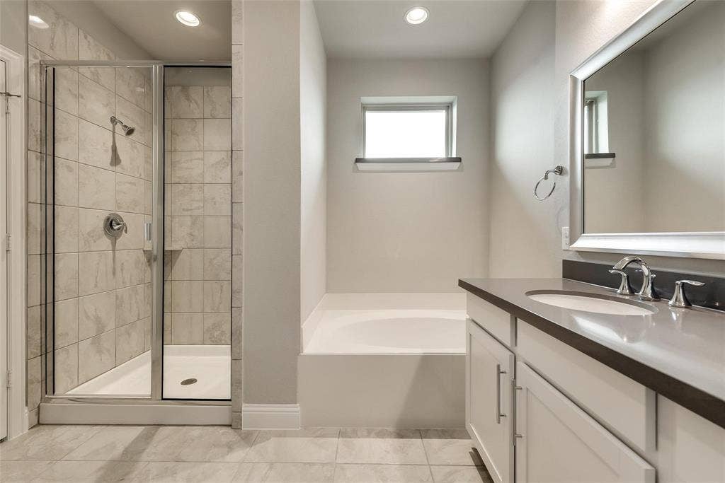 Shower with glass enclosure, separate soaking tub, double vanity with integrated sinks, framed mirror, and tiled flooring