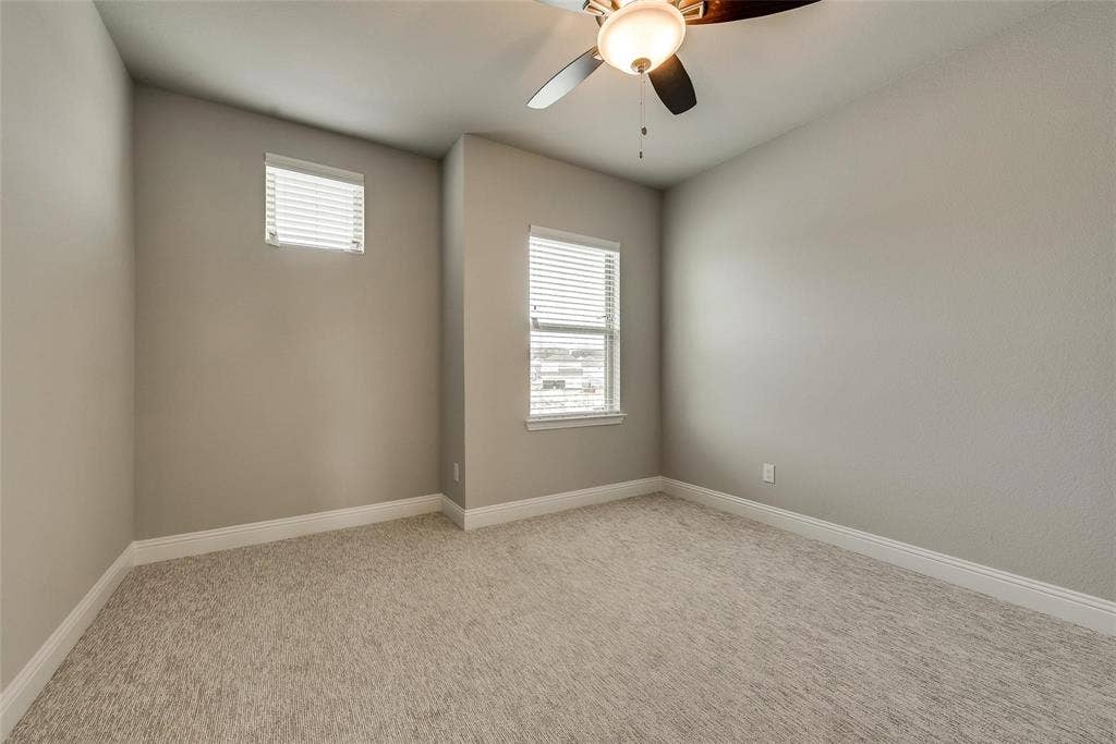 Bright secondary bedroom featuring neutral painted walls, plush carpeting, dual windows with plantation shutters, and ceiling fan with integrated lighting