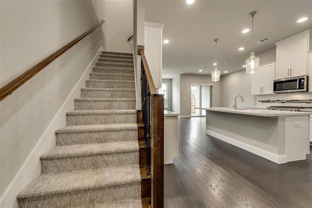 Carpeted staircase with wood handrail, wood-finish flooring, recessed lighting, kitchen island with sink, and stainless steel microwave