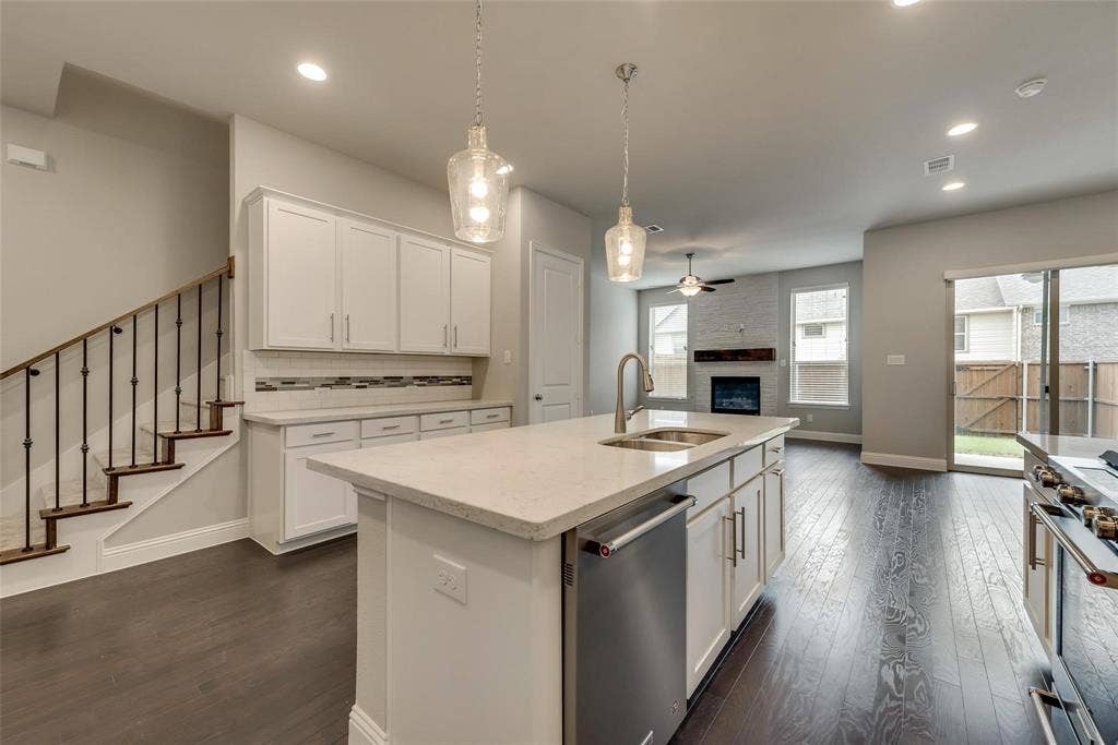 Wood-finish flooring throughout, kitchen island with integrated sink, stainless steel appliances, white cabinetry with modern hardware, and a fireplace with a stone surround