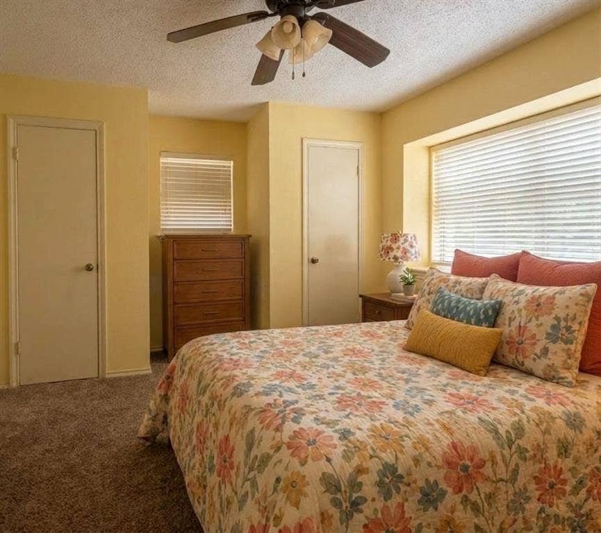 Staged AI furniture;  ceiling fan with lighting, window with horizontal blinds, and two solid panel doors to closets.