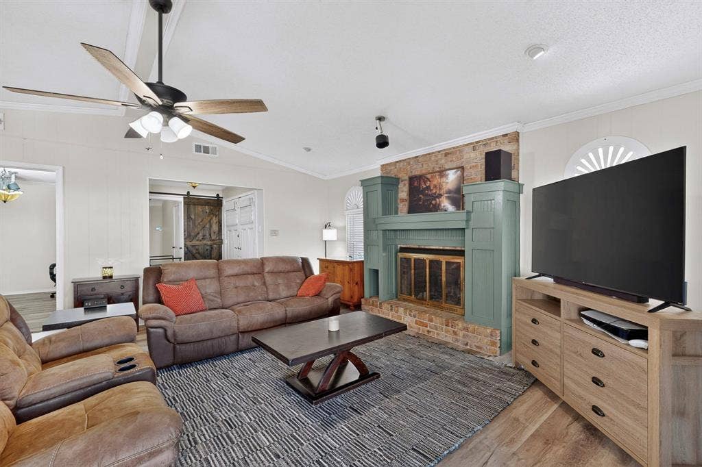 1418 Stony Brook Lane, Garland, TX 75043 Vaulted ceiling with a ceiling fan, wood-finish flooring, brick-faced fireplace with a painted mantel, decorative window accent, and a barn door entry