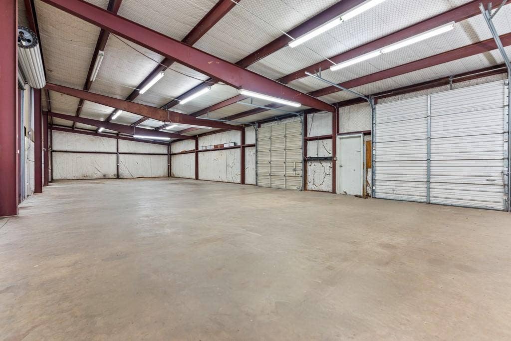 414 E Marshall Street, Gilmer, TX 75644 Spacious interior featuring a concrete floor, exposed metal beam construction, multiple overhead lighting fixtures, and several roll-up doors