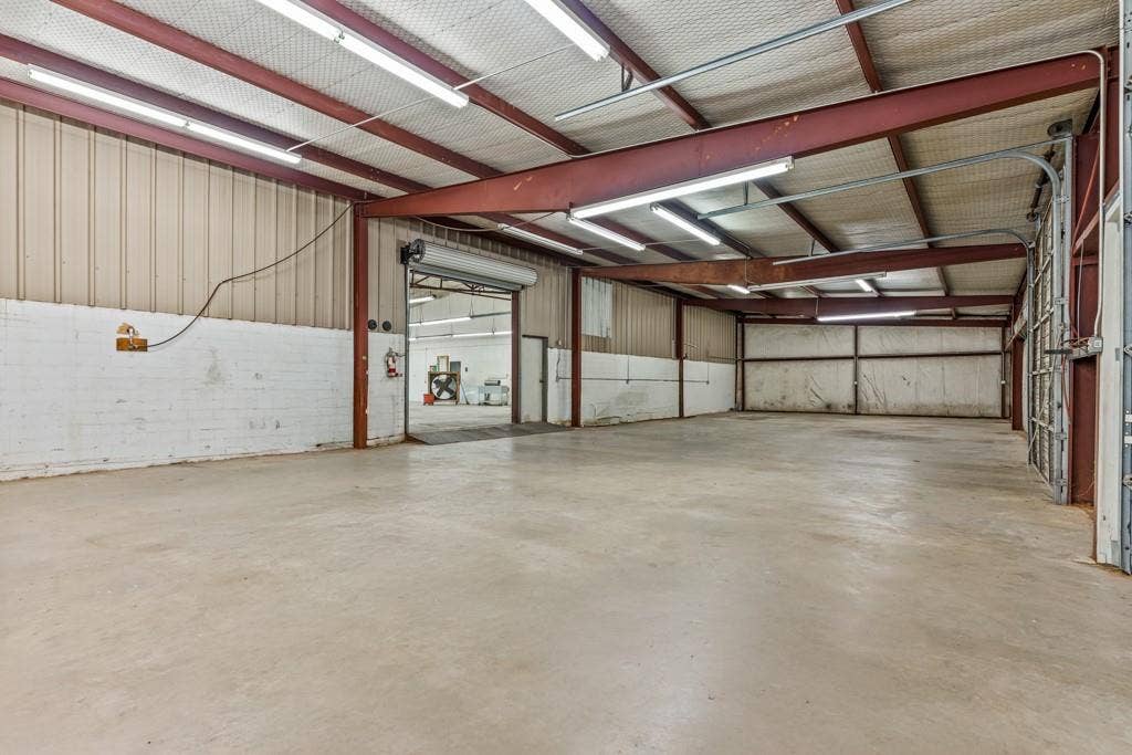 414 E Marshall Street, Gilmer, TX 75644 Spacious warehouse featuring a concrete floor, exposed structural beams, corrugated metal walls, overhead fluorescent lighting, and two roll-up doors