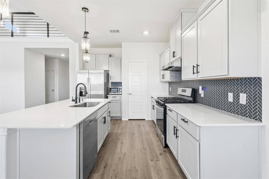 1536 Estivella Drive, Little Elm, TX 75068 Modern kitchen featuring white cabinetry, a large kitchen island with an integrated sink, stainless steel appliances, wood-finish flooring, and a dark herringbone tile backsplash