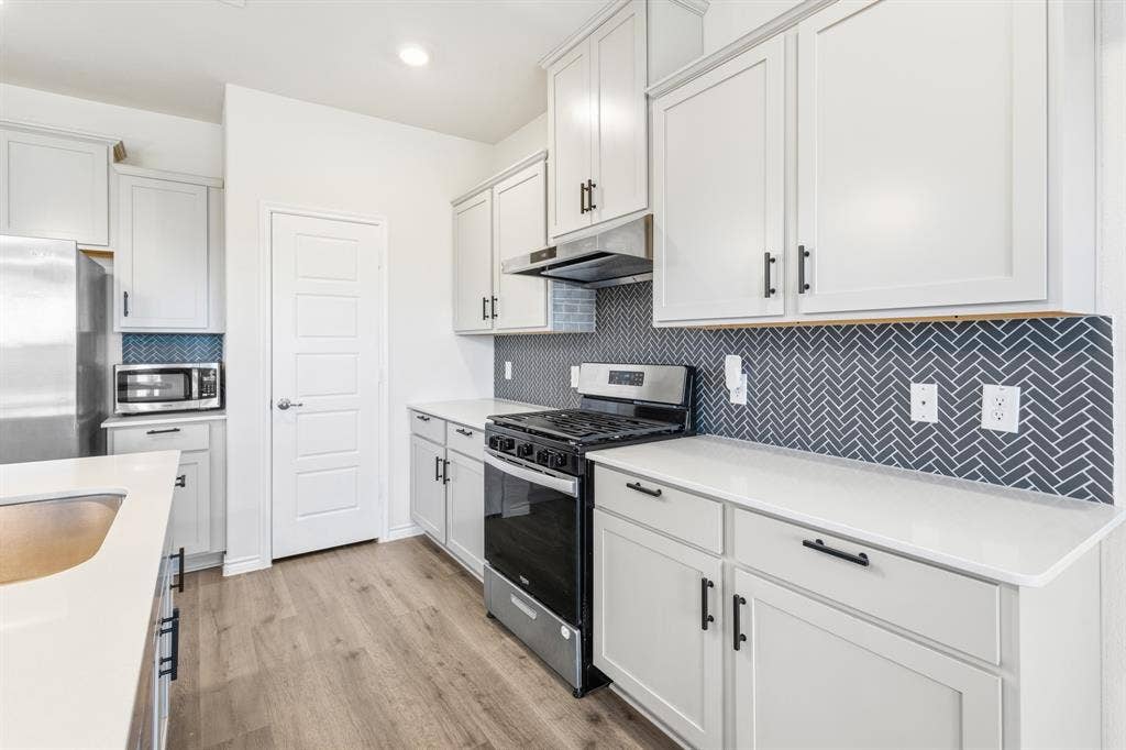 1536 Estivella Drive, Little Elm, TX 75068 Contemporary kitchen featuring light wood-finish flooring, white cabinetry with black hardware, white countertops, a herringbone tile backsplash, and stainless steel appliances