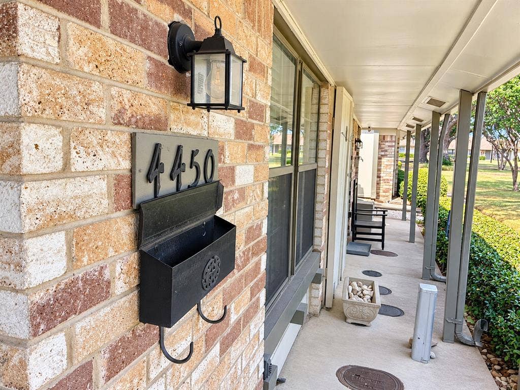Covered front porch featuring a brick exterior facade, mounted metal mailbox with house numbering, and an exterior wall-mounted carriage light fixture