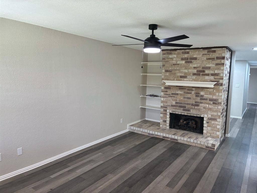Brick fireplace with a mantel, integrated wall shelving, wood-finish flooring, and a ceiling-mounted light fixture with fan
