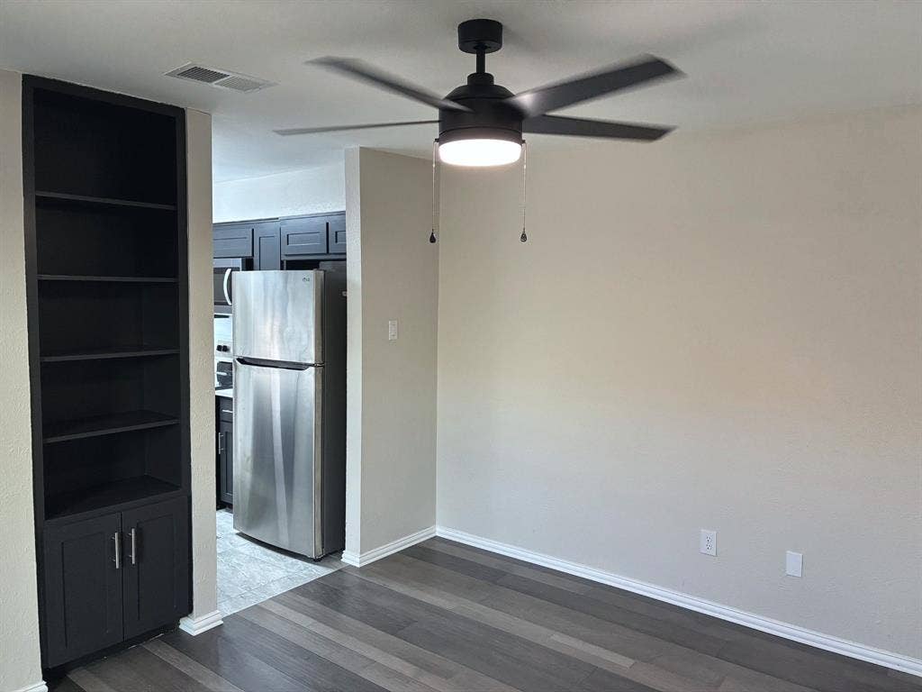 Matte black ceiling fan with integrated lighting, custom dark-toned built-in cabinetry, stainless steel refrigerator, and wood-finish flooring