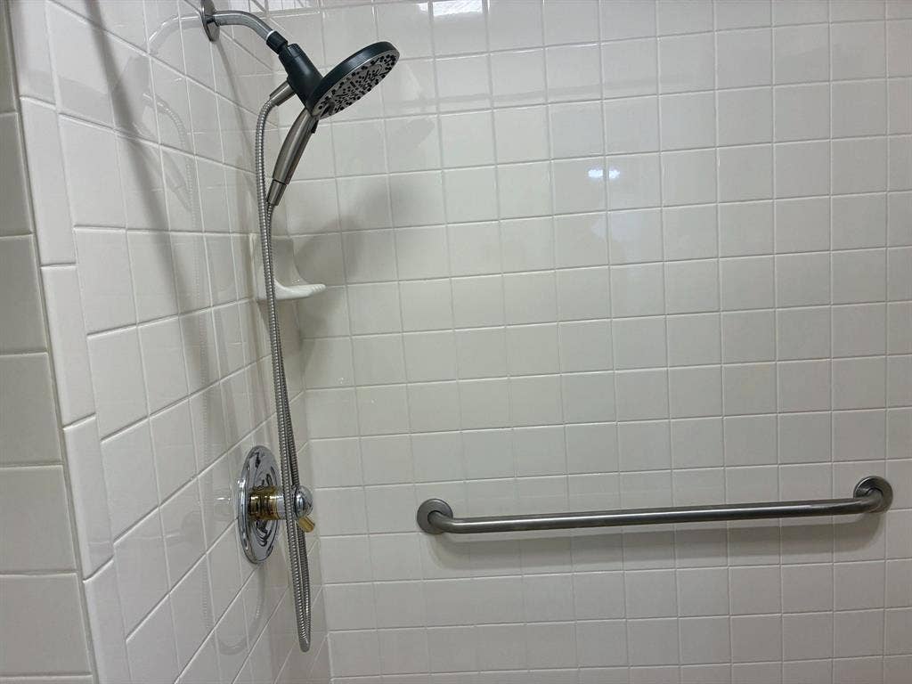Shower enclosure featuring white square wall tile, a handheld showerhead, a built-in recessed soap dish, and a horizontal metal grab bar