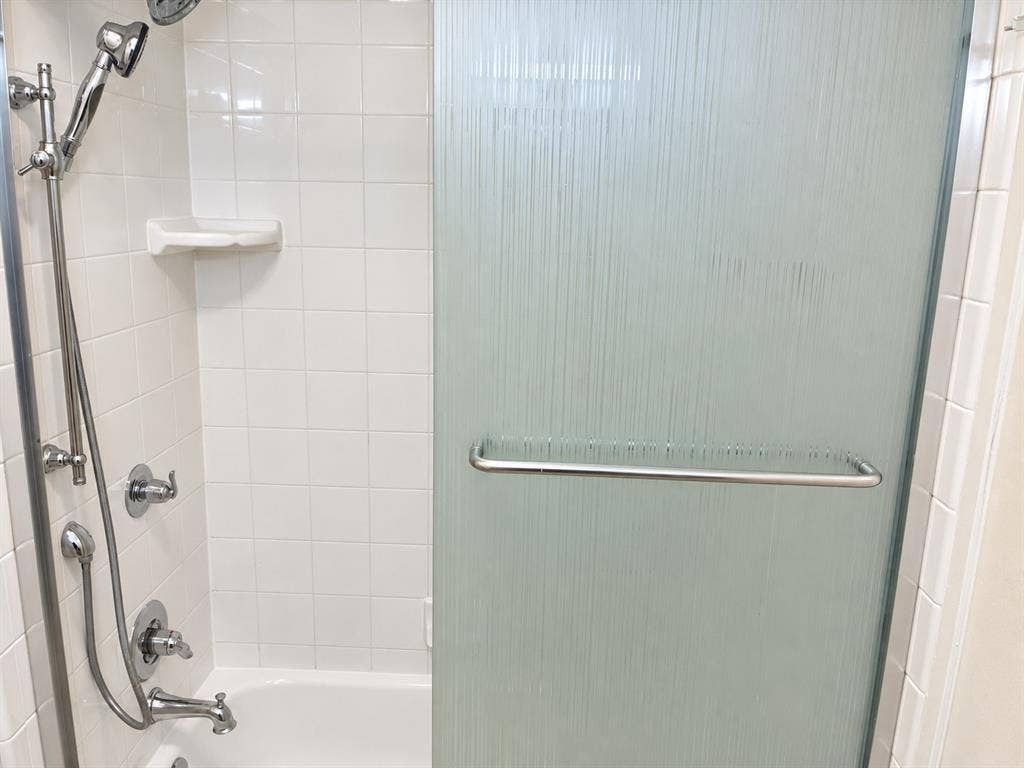 Shower enclosure featuring a chrome handheld shower head, integrated wall-mounted soap dish, textured glass sliding door with a horizontal chrome handle, and white tiled surround