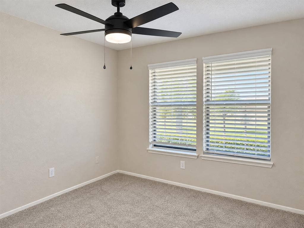 Neutral wall paint, plush carpeting, dual windows with fitted blinds, and a contemporary matte black ceiling fan with integrated lighting