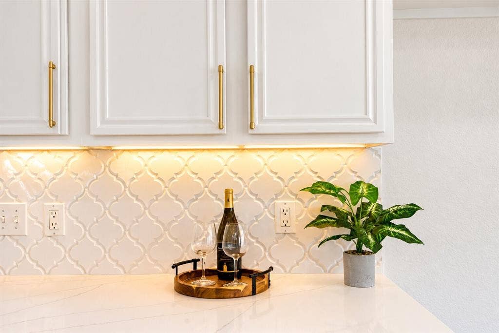 White cabinetry with gold-tone hardware, under-cabinet lighting, arabesque tile backsplash, and a solid surface countertop