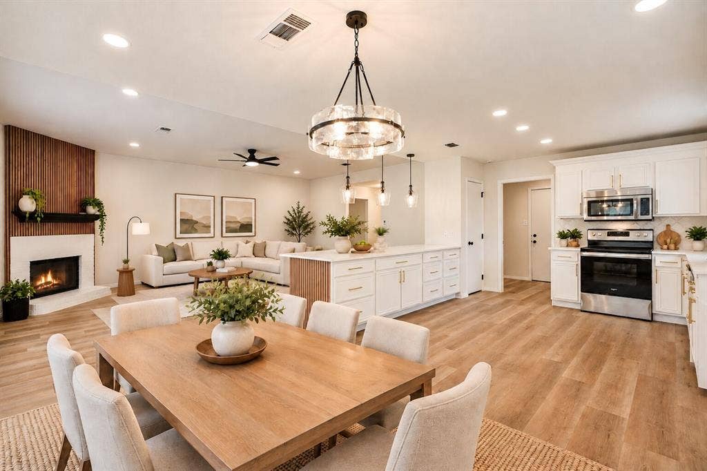 Open-concept living space featuring wood-finish flooring, a contemporary fireplace with a wood-paneled accent wall, a kitchen island with storage, and a dining area illuminated by a modern chandelier