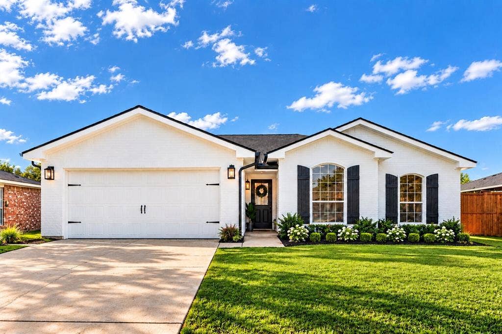 White brick exterior with black trim, an attached garage, arched windows with shutters, and manicured landscaping