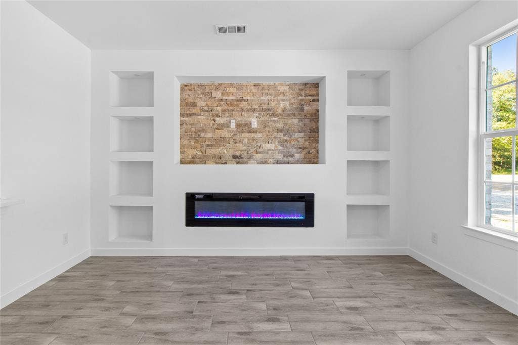 7520 Double Bridge Road, Mabank, TX 75156 Modern entertainment space featuring custom built-in shelving flanking a brick accent wall, linear electric fireplace with blue LED accent lighting, wood-finish plank flooring, and abundant natural light from large picture window