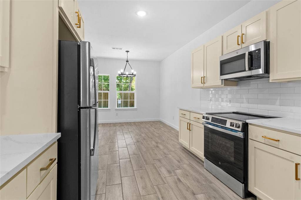 7520 Double Bridge Road, Mabank, TX 75156 Galley-style kitchen featuring cream-colored cabinetry with brass hardware, stainless steel appliances including a French-door refrigerator and built-in microwave, subway tile backsplash, wood-finish flooring, and abundant natural light from dual windows