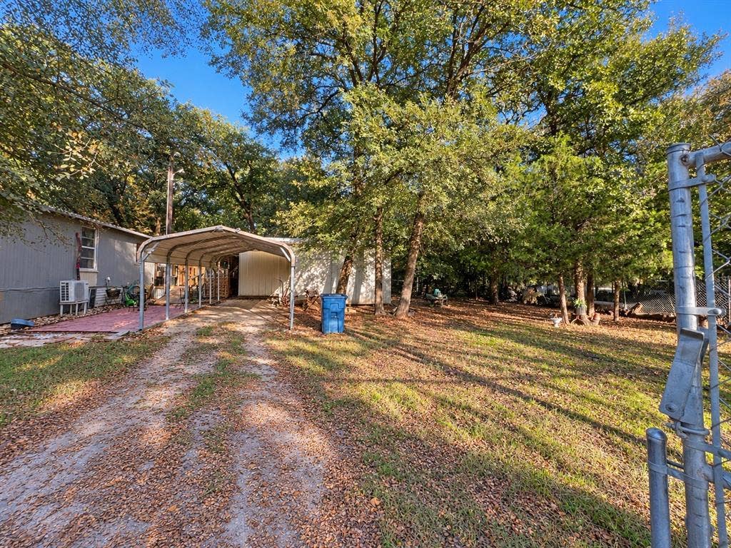 632 Cumberland Trail, Valley View, TX 76272 Gravel driveway leading to a metal carport structure, a side patio with brick pavers, and a spacious grassy area with mature trees