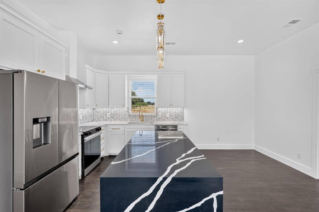 102 Shadywood Road, Trinidad, TX 75163 Kitchen featuring a large central island with a dramatic veined countertop, stainless steel appliances, white cabinetry with gold-finish hardware, and a herringbone tile backsplash
