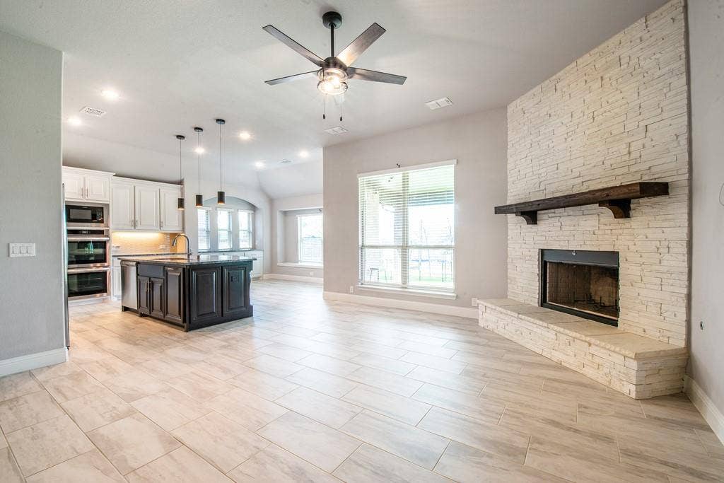 Open-concept living area featuring wood-finish flooring, a stone-clad fireplace with a dark wood mantel, and an island kitchen with a dark base and light upper cabinetry