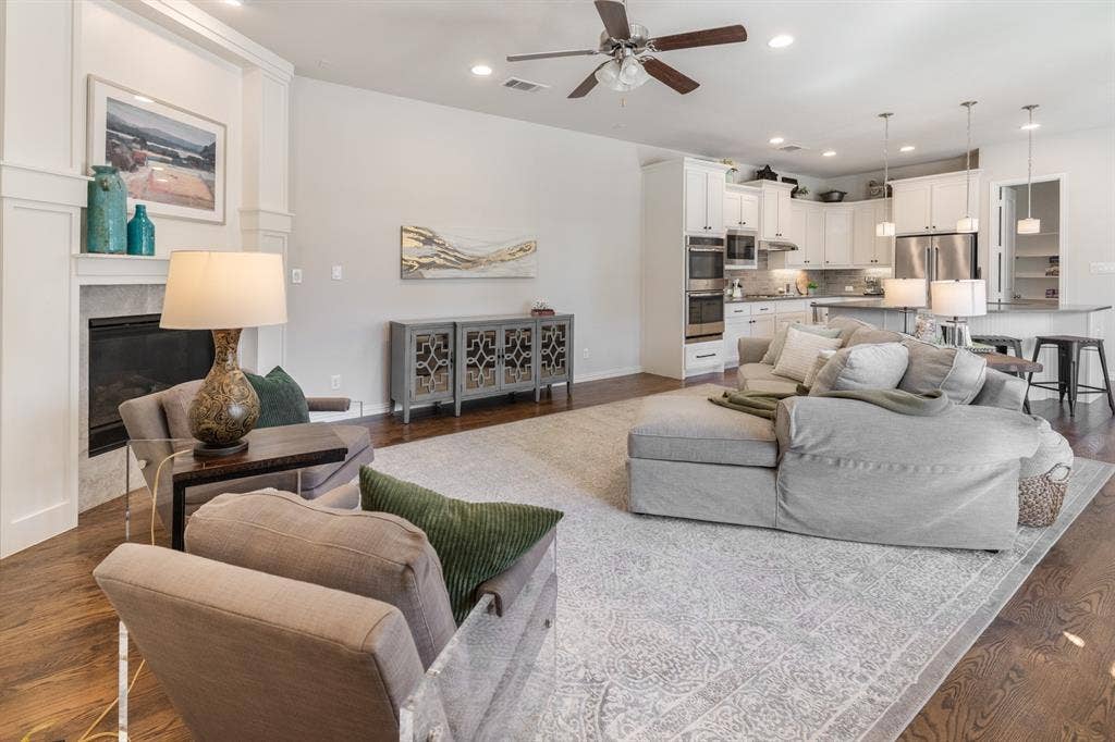 Hardwood-finish flooring throughout, a ceiling fan with integrated lighting, recessed lighting, a built-in fireplace with a stone-surround, and a kitchen featuring white cabinetry and stainless-steel appliances
