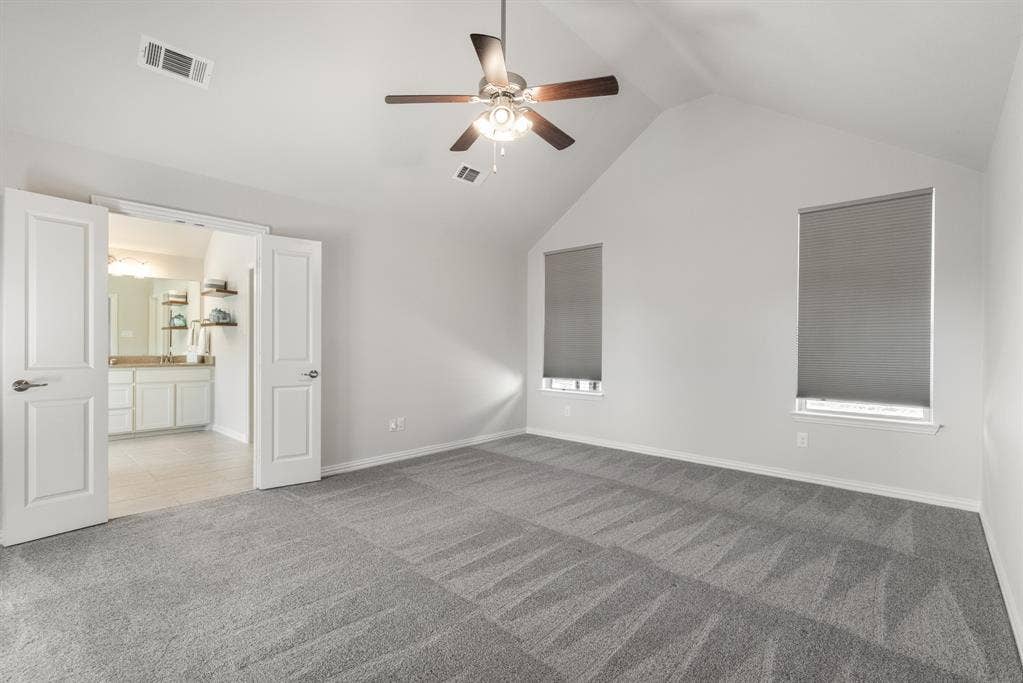 Vaulted ceiling, ceiling fan, two windows with roller shades, gray carpeting, and double doors opening to a bathroom