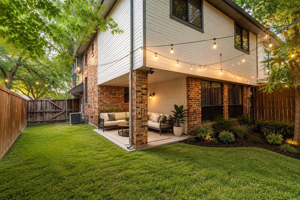 Expansive backyard featuring a manicured lawn, a covered patio with brick pillars, and mature trees providing natural shade