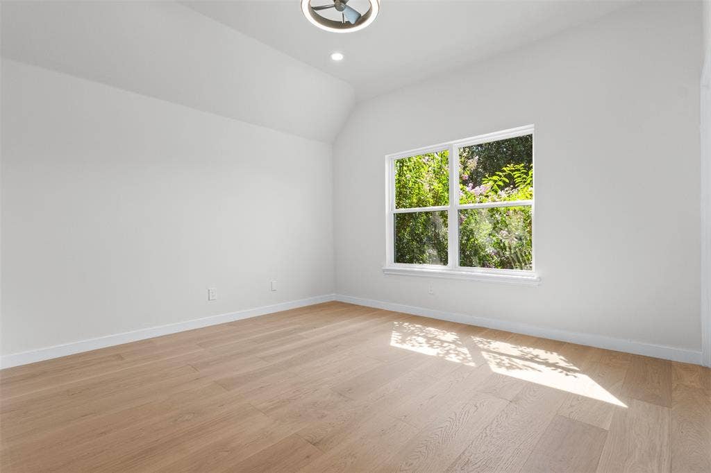 Vaulted ceiling with a flush-mount light fixture, recessed lighting, wood-finish flooring, and a large window with exterior greenery