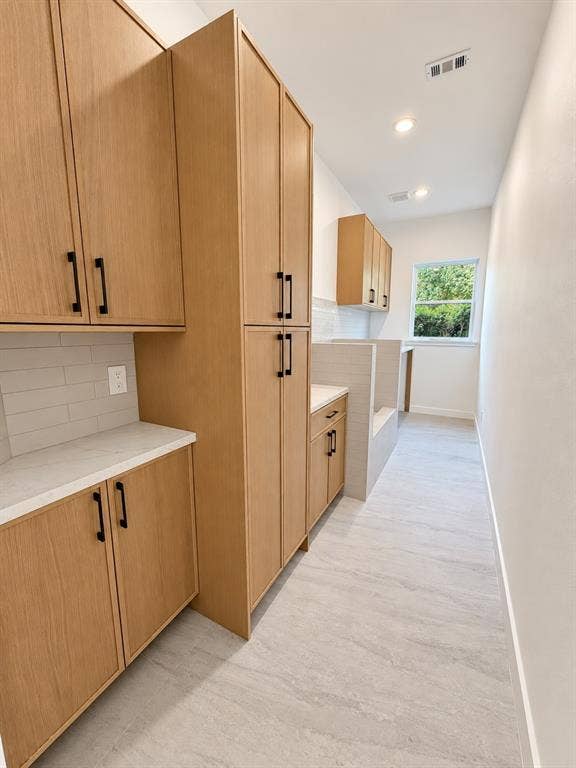 Light wood-finish cabinetry with black hardware, white countertops, tile backsplash, and light gray tile flooring