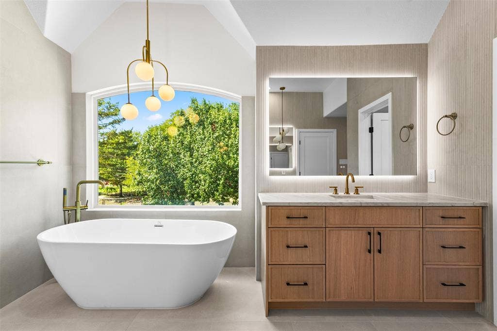 Vaulted ceiling bathroom featuring a freestanding soaking tub, wood-finish vanity with ample storage, and an illuminated mirror