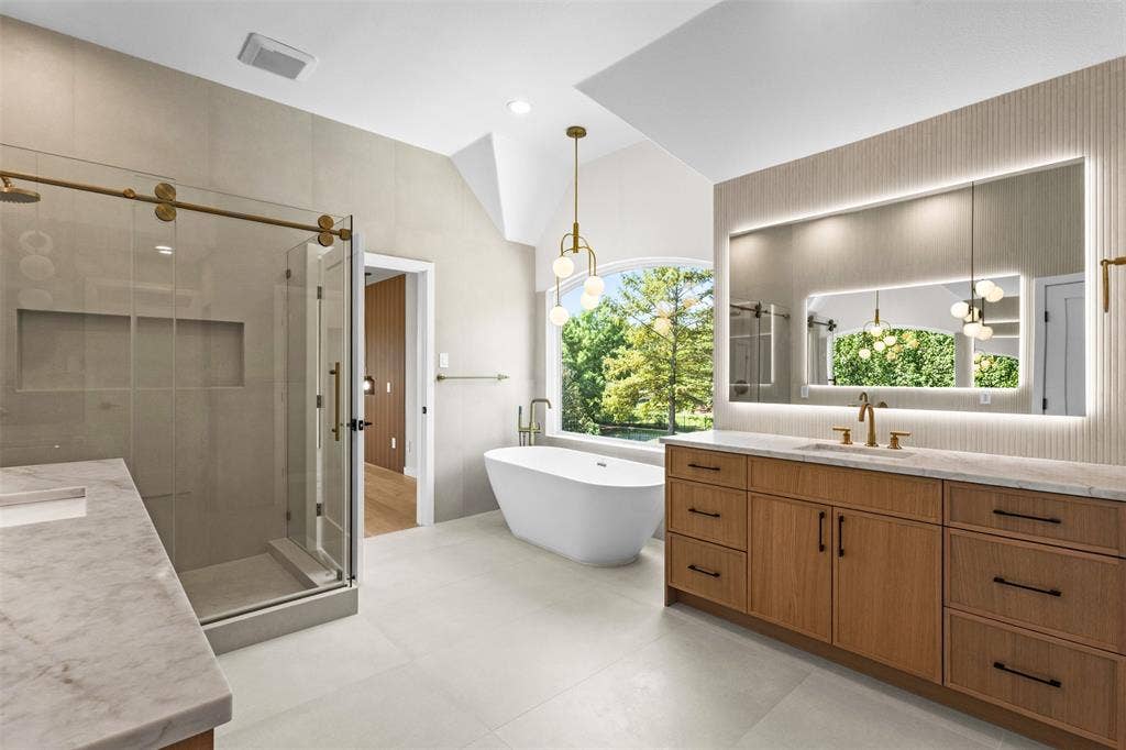Freestanding soaking tub, dual vanity with wood-finish cabinetry, illuminated medicine cabinet, walk-in shower with glass enclosure, and vaulted ceiling