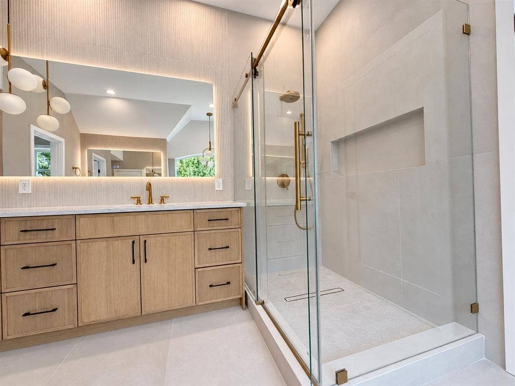 Bathroom featuring a wood-finish vanity with a light-colored countertop, an illuminated mirror, a glass-enclosed shower with gold-tone fixtures, and a tiled shower niche