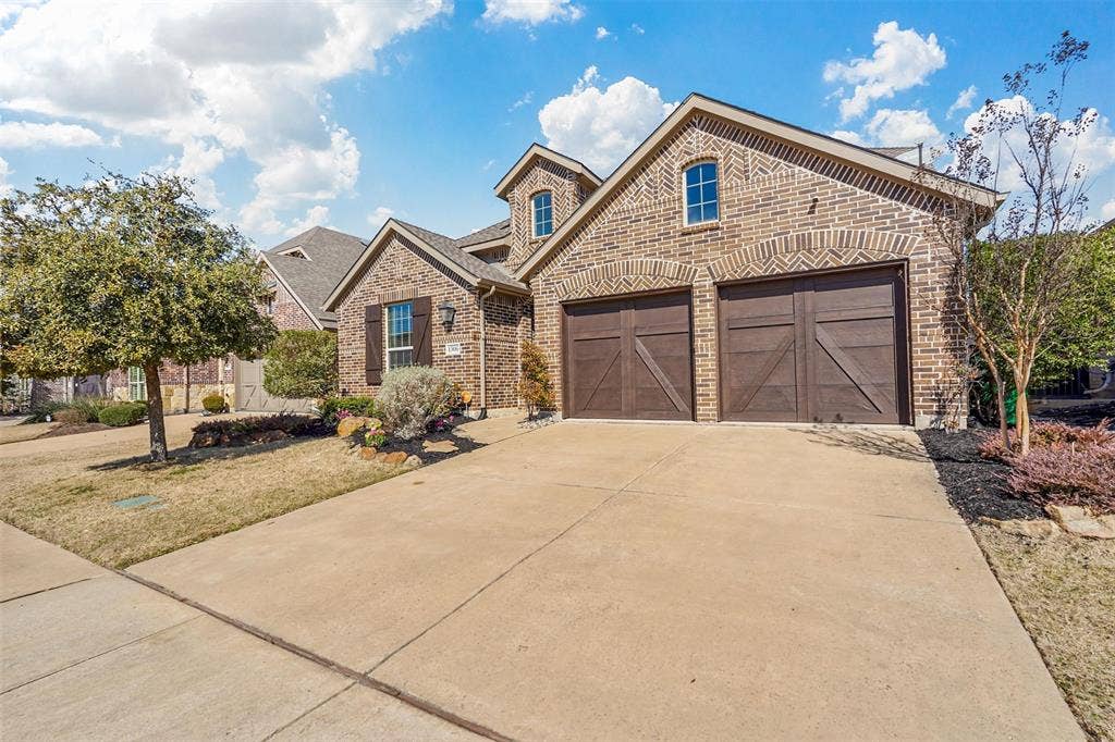 1506 Carnation Street, Celina, TX 75078 Brick exterior with arched window details, attached two-car garage with wood-finish doors, concrete driveway, and landscaped front yard with mature trees