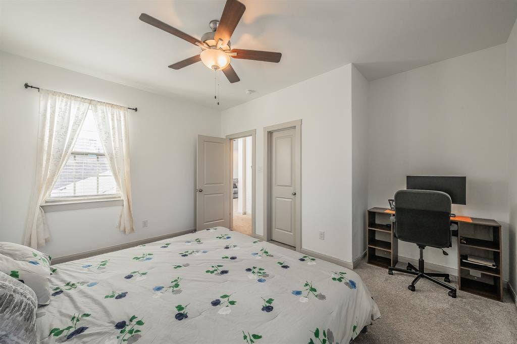 First of two second floor bedrooms with carpeted flooring, a window with trim, a ceiling fan with integrated lighting, and spacious closet with extra room for a desk or armoire
