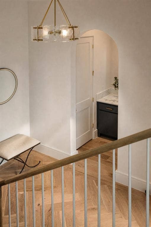 Herringbone wood-finish flooring, staircase with natural wood handrail and white spindles, arched doorway leading to a built-in cabinet with stone countertop, and a brass and glass chandelier